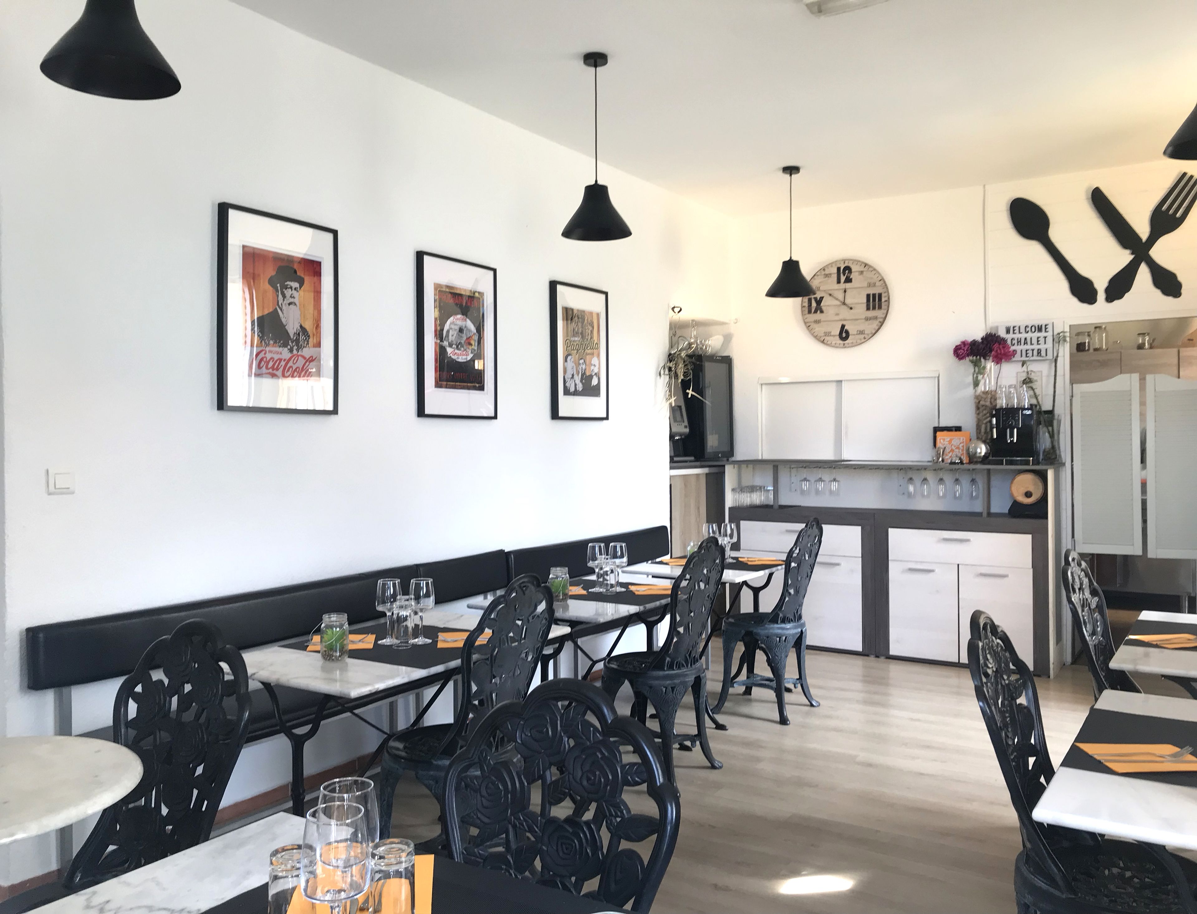 Un restaurant avec des chaises en métal noir, des tables blanches et des lampes suspendues noires. Les murs sont ornés de tableaux encadrés et il y a une horloge.