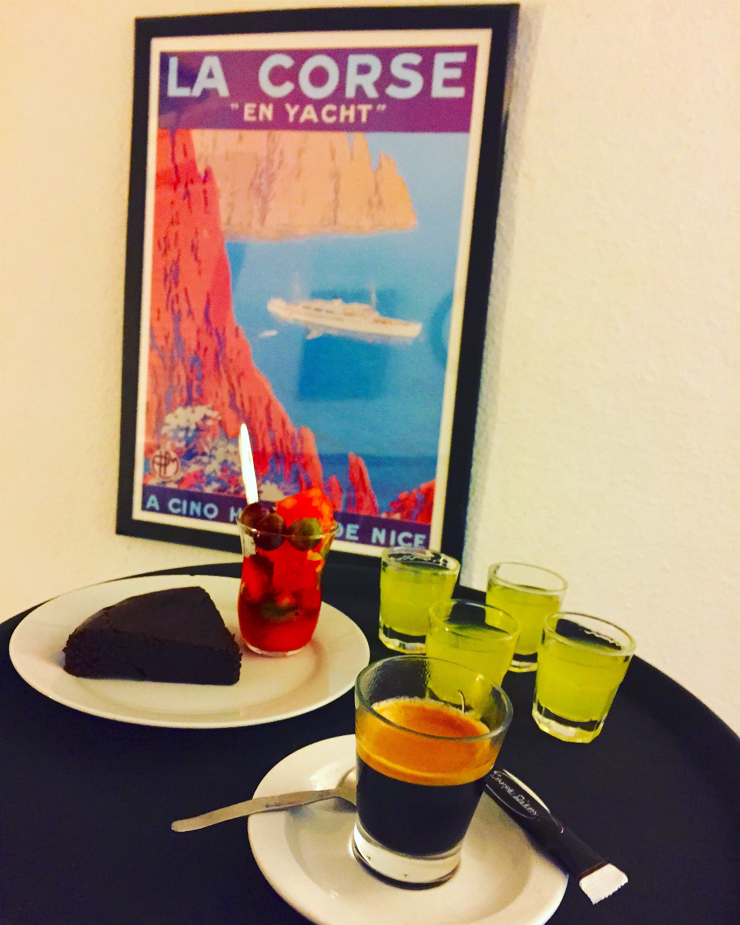 A table with a slice of cake, a drink with a straw, and shot glasses with a yellow liquid. A framed poster with the text "LA CORSE EN YACHT" is visible in the background.