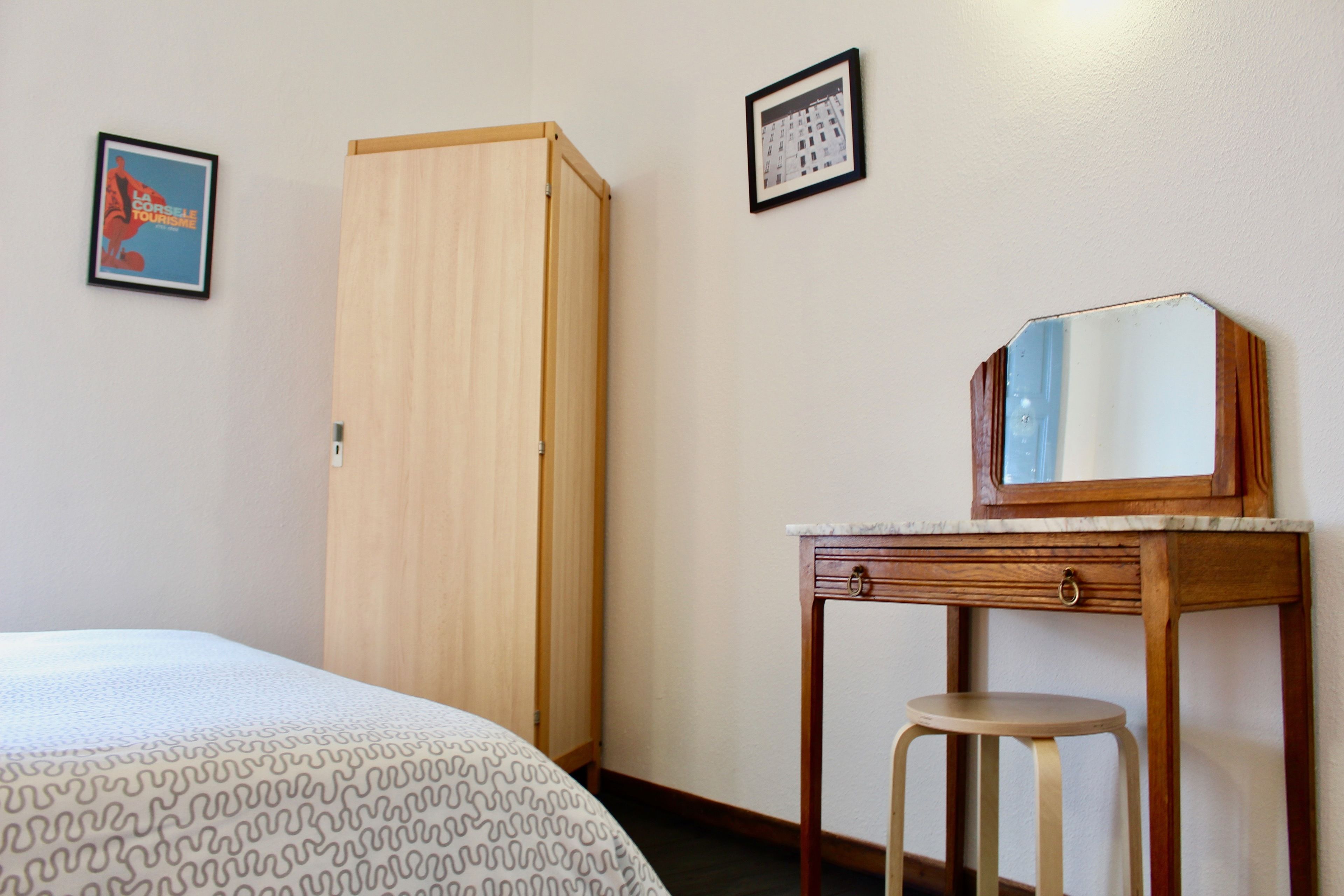 A hotel room with a bed, a wooden wardrobe, a small table with a mirror, and two framed pictures on the wall.