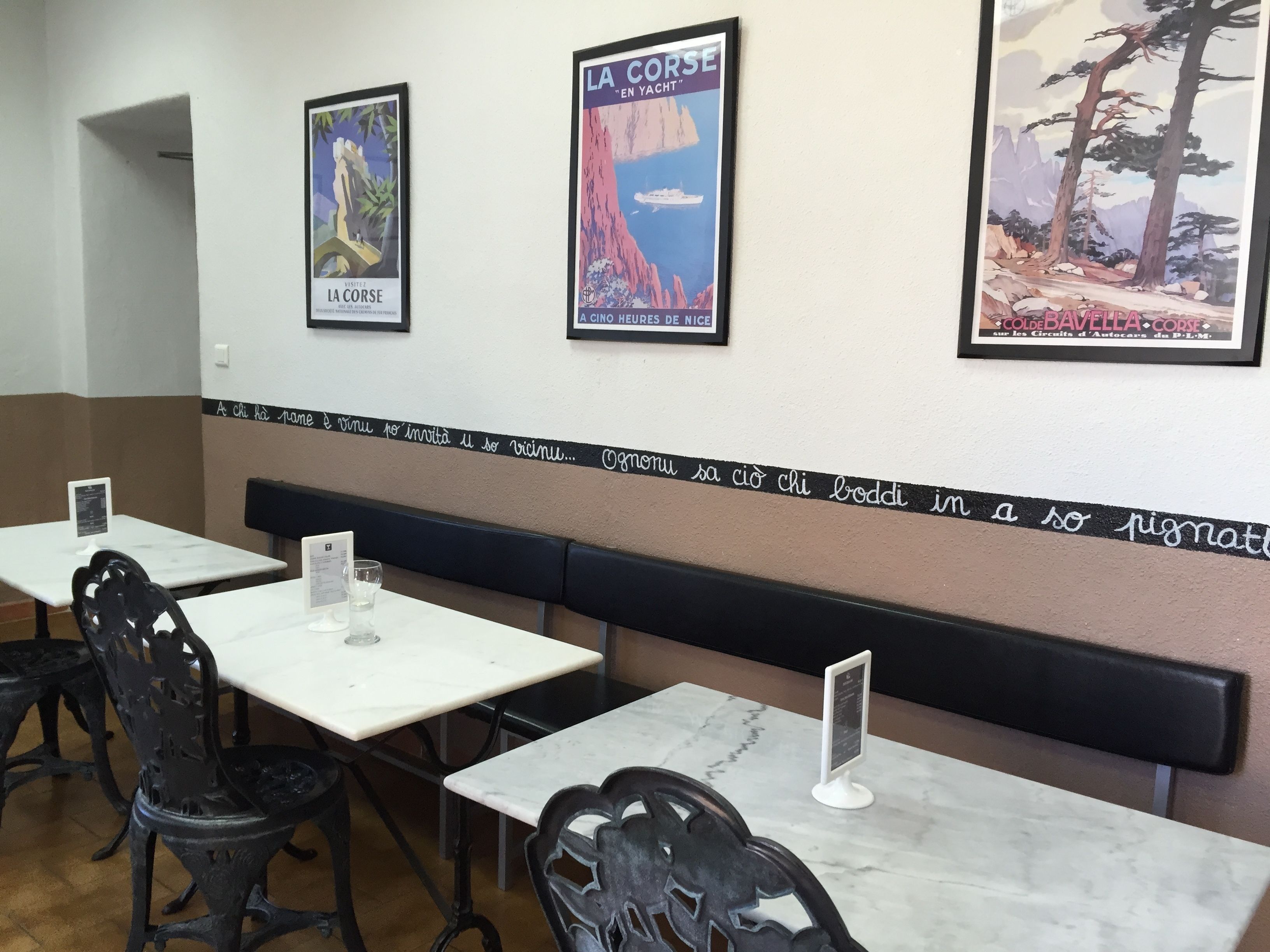 A restaurant interior with white marble tables and black metal chairs. There are framed posters on the wall, one of which reads "LA CORSE EN YACHT".