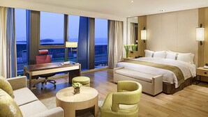 A hotel room with a large bed, a desk, a chair, a round coffee table, and a view of the cityscape through large windows.