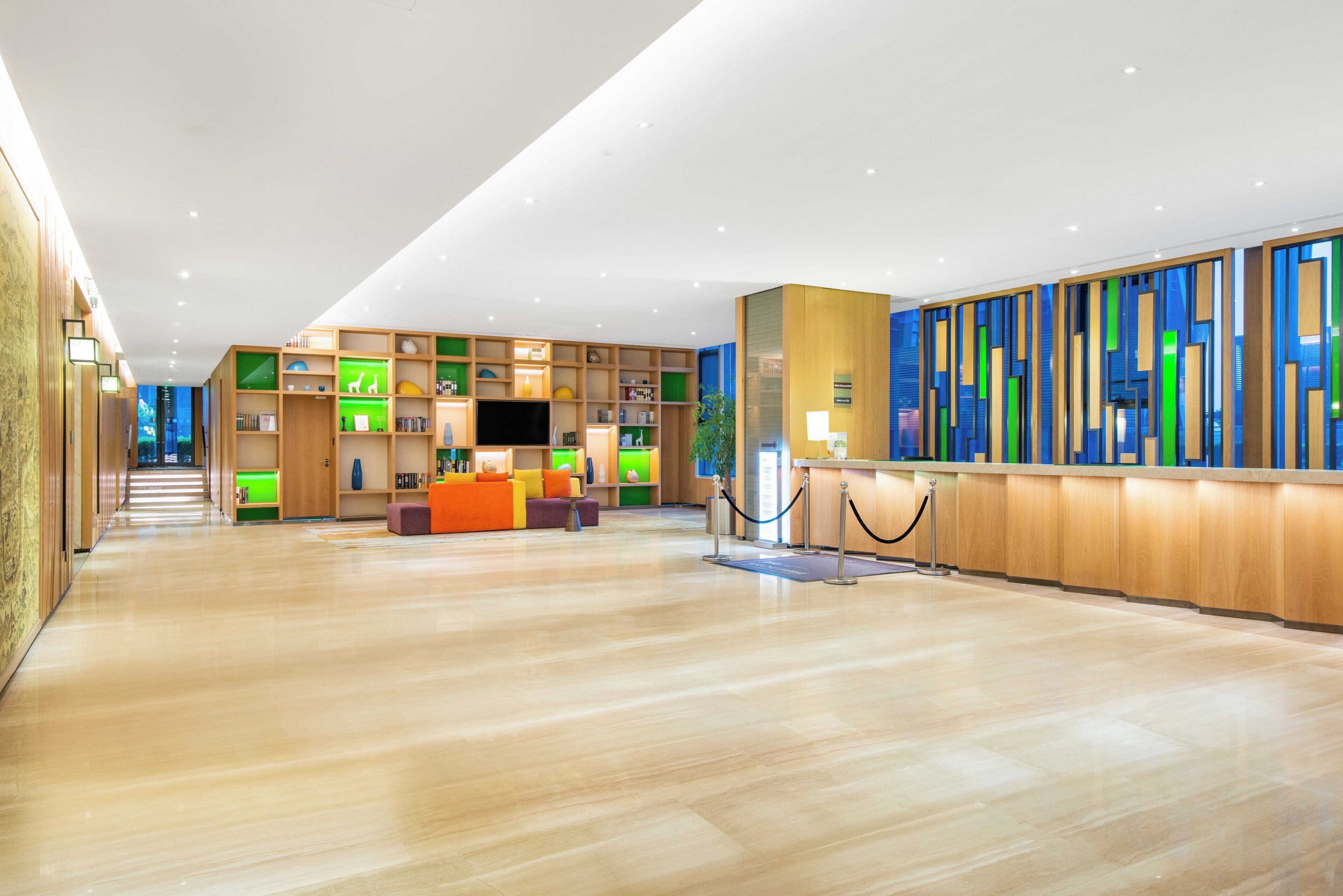 A spacious lobby with a wooden reception desk, a seating area with colorful cushions, and a large bookshelf filled with various items.