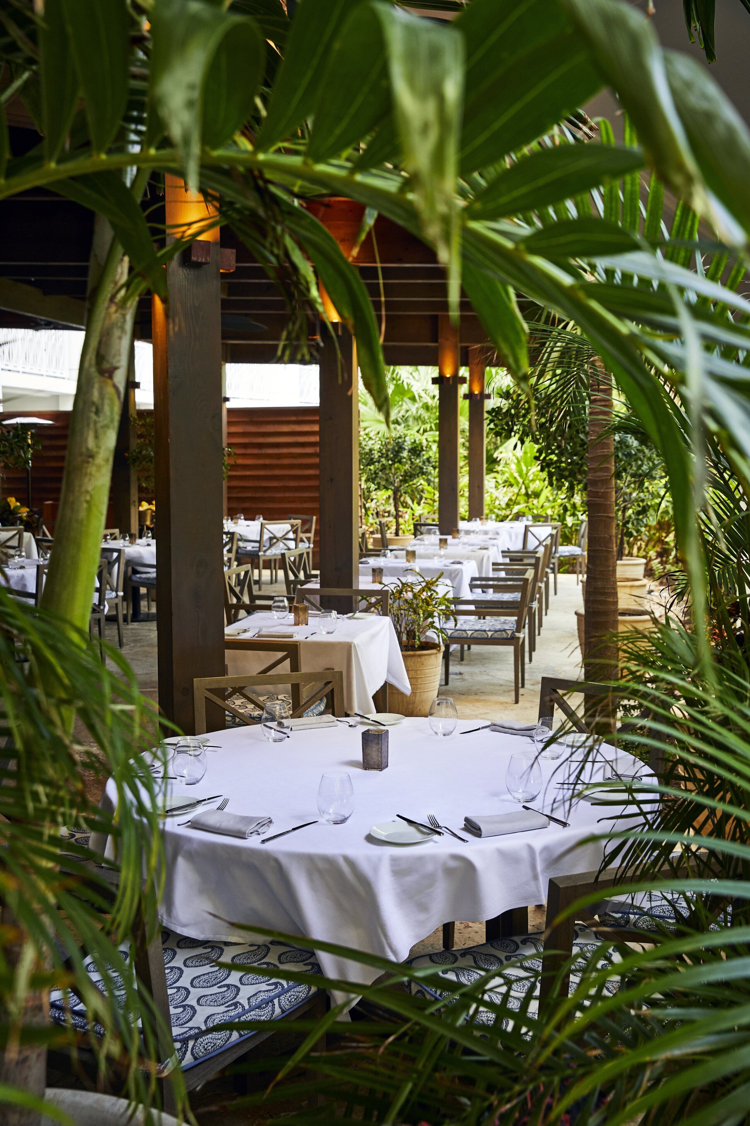 An outdoor dining area with tables set for two, surrounded by lush greenery and wooden pillars.