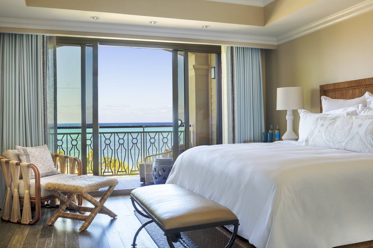 Een hotelkamer met een groot bed, een sofa, een bankje en uitzicht op zee vanaf een balkon.