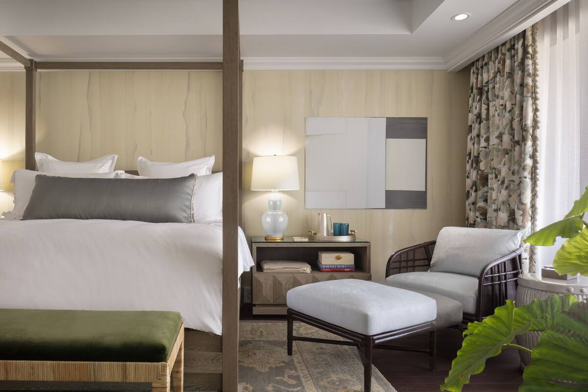 Een hotelkamer met een bed, nachtkastje, stoel, poef en een grote plant.