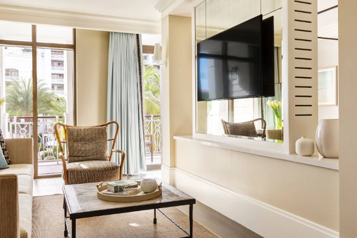 Een woonkamer met een groot raam, een flatscreen televisie, een rieten fauteuil, een bijzettafel met boeken en een balkon met uitzicht op palmbomen.