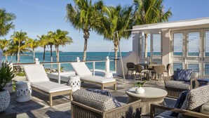 A sunlit outdoor lounge area with lounge chairs, wicker furniture, and a view of the ocean.