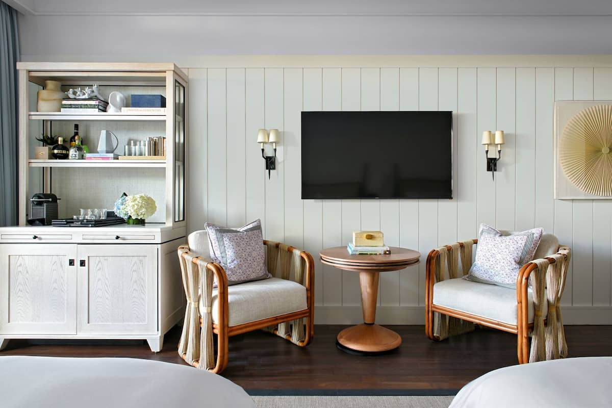 A hotel room with a bed, two wicker chairs, a small round table, a minibar, a flat-screen TV, and a wall-mounted lamp.