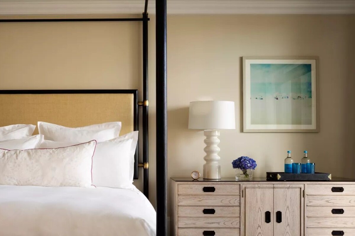 A neatly made bed with white linens and a headboard, a bedside table with a lamp, a framed artwork on the wall, and a small clock.