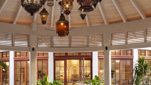 A spacious indoor area with wooden ceiling beams, hanging lanterns, a central bar area with stools, and large windows allowing natural light.