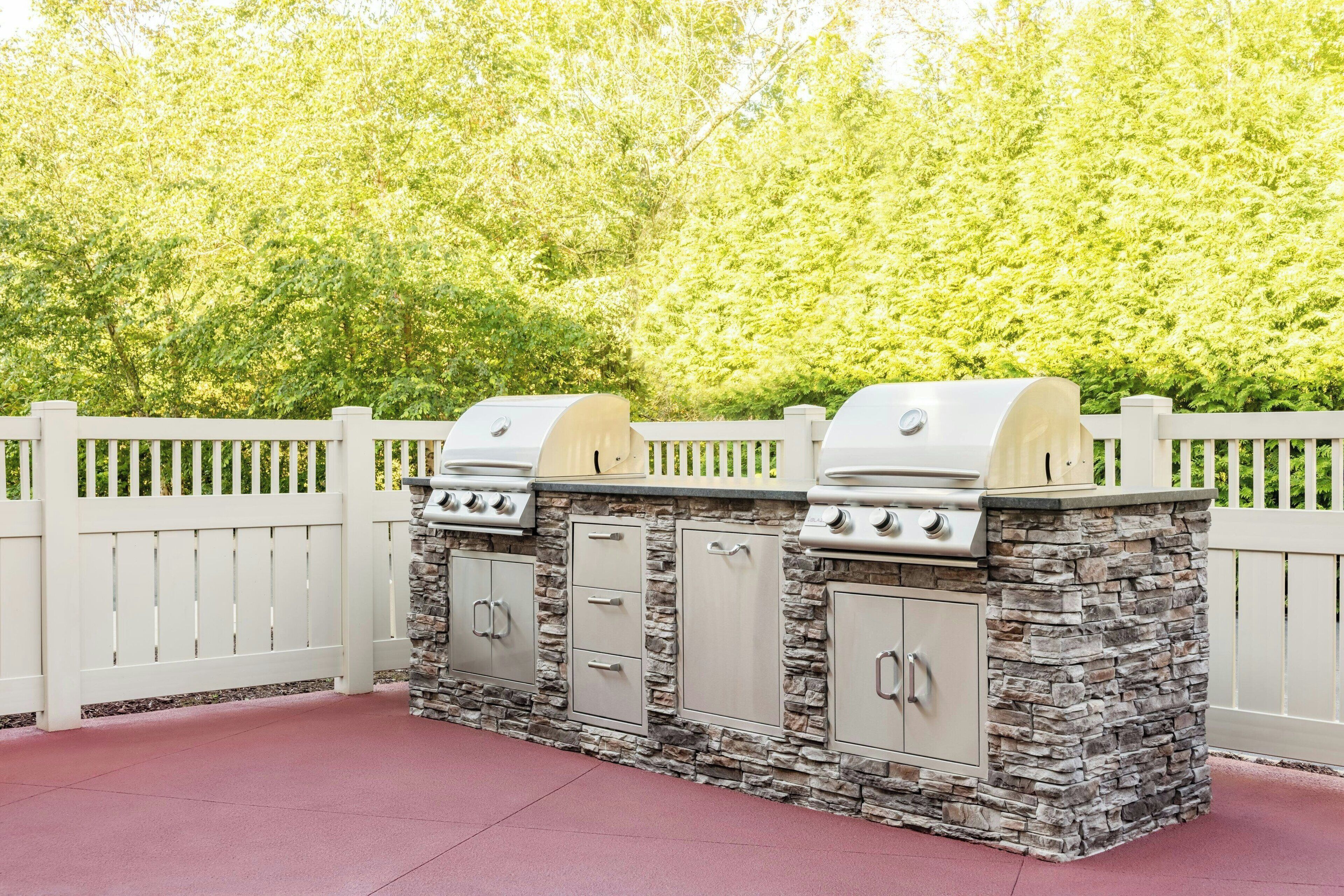 Un barbecue est installé contre une clôture blanche en bois, dont la base est en pierre, et est entouré d’une végétation luxuriante.