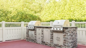 Grill med grillkul foran et hvidt plankeværk med stensokkel, omgivet af frodig vegetation.