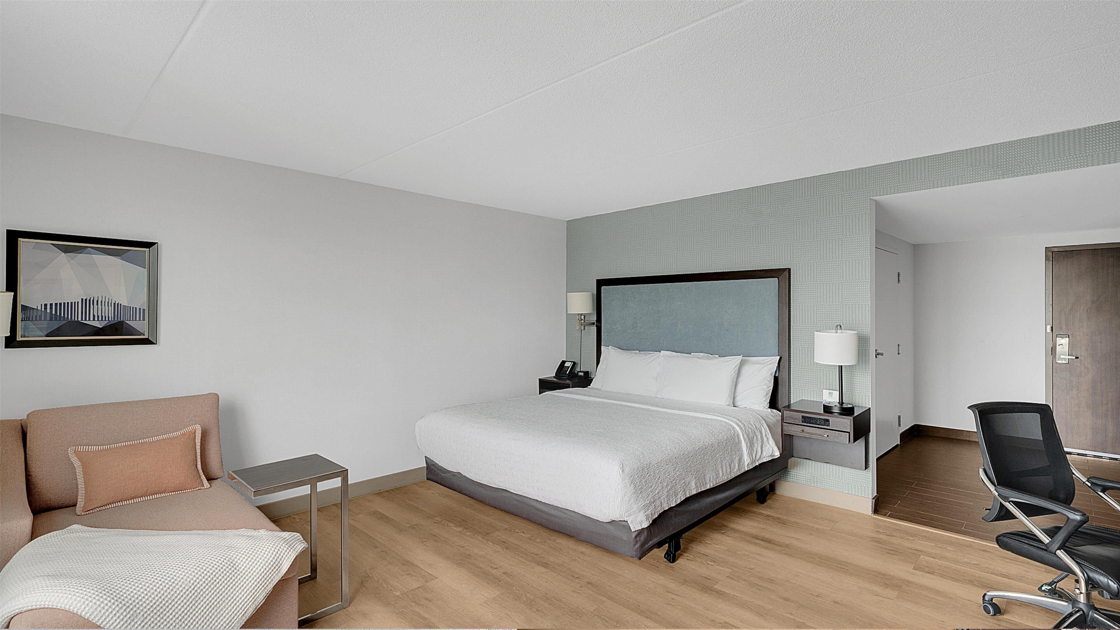 A modern hotel room with a large bed, bedside tables, a desk, a chair, and a framed picture on the wall.