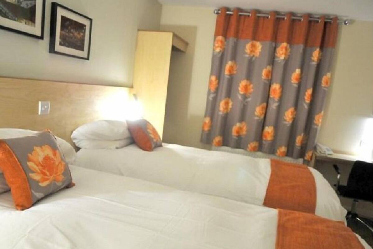 A hotel room with two beds, a nightstand, a lamp, and a window with floral curtains.