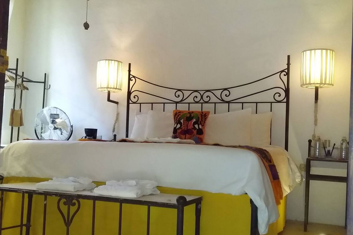 Ein Bett mit Metallgestell, zwei Nachttischlampen, ein Nachttisch mit Flaschen und ein Wandventilator.