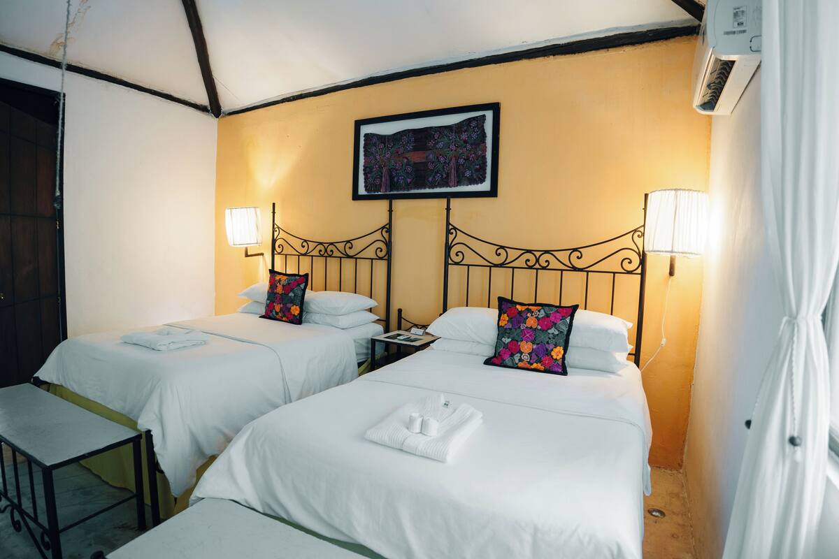 Ein Hotelzimmer mit zwei Betten, jeweils mit weißen Laken und dekorativen Kissen. Es gibt einen kleinen Tisch mit einem Stuhl, eine Wand-Klimaanlage und ein gerahmtes Bild über den Betten.