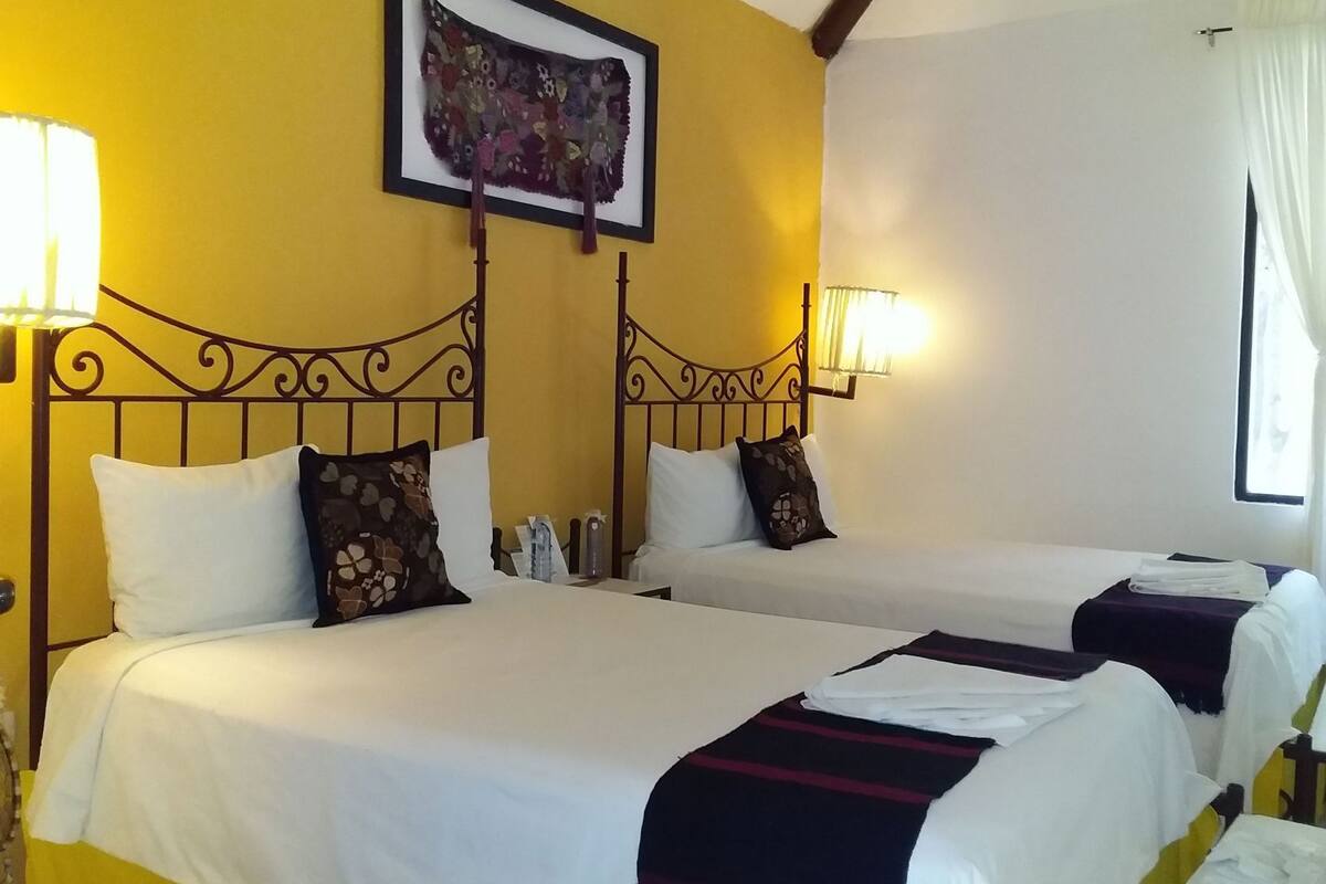 Ein Hotelzimmer mit zwei Betten, jeweils mit weißer Bettwäsche und dekorativen Kissen. Darüber hängt ein gerahmtes Kunstwerk, und eine Wand im Zimmer ist gelb gestrichen.