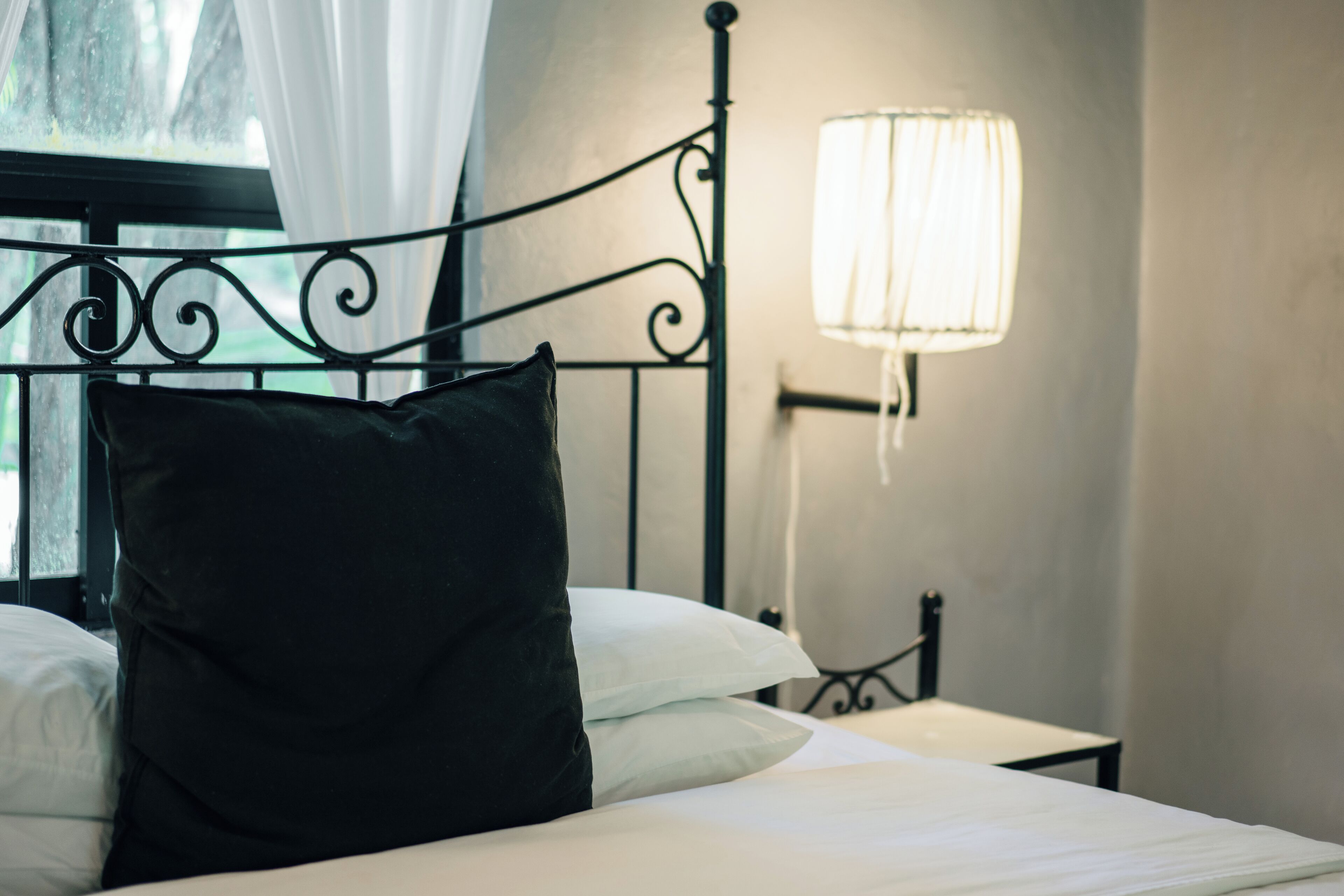 Una cama bien hecha con un almohadón negro y uno blanco, un cabecero de metal, una mesita de noche con una lámpara y una ventana con cortinas.