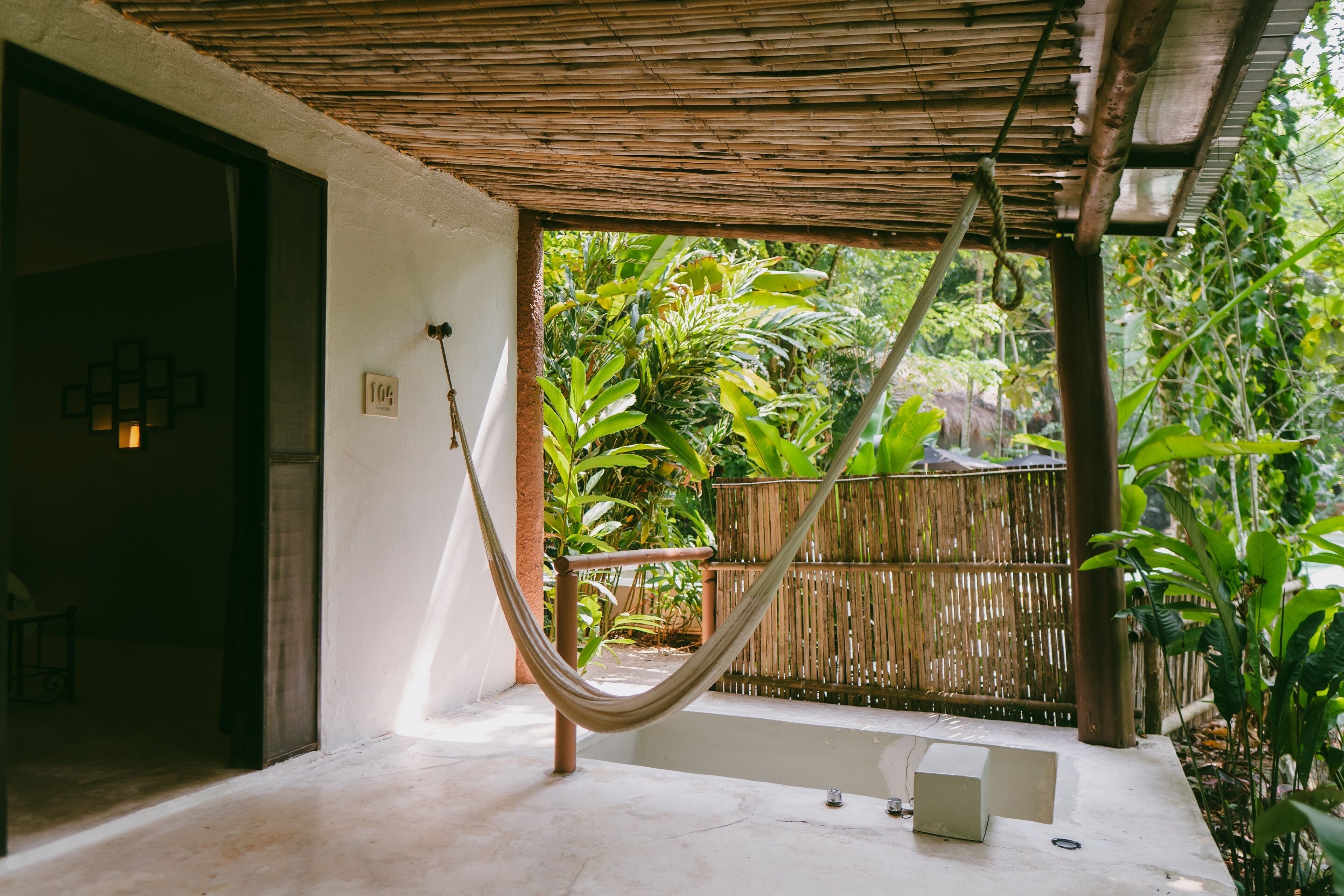 A hammock under a bamboo roof, a small pool, and lush greenery in the background.