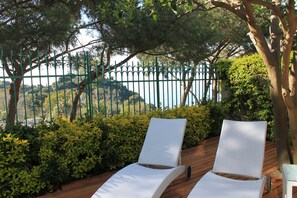 Zwei weiße Lounge-Stühle auf einer Holzterrasse mit Blick auf einen grünen Zaun und Bäume.