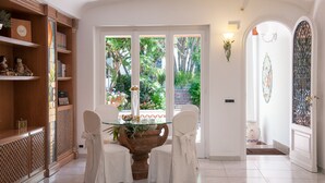 A dining area with a glass-top table, white chairs, and a view of a garden through a glass door.