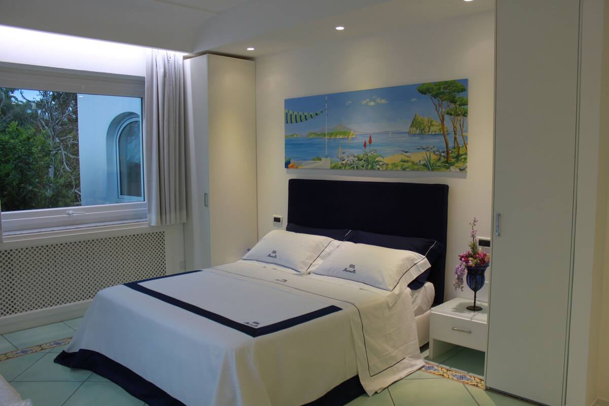 Une chambre à coucher avec un lit, une table de chevet, un tableau et une fenêtre avec des rideaux.