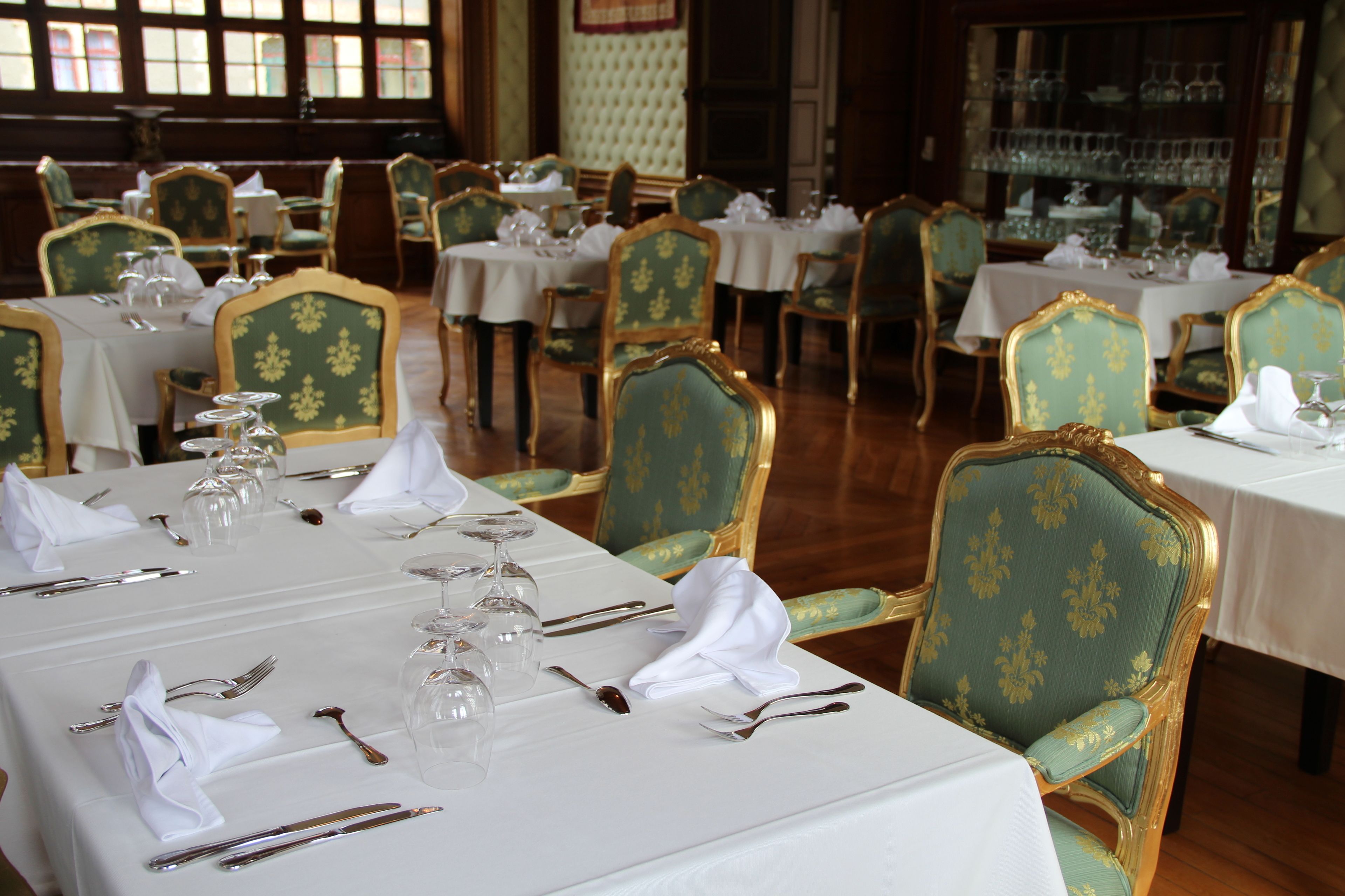 Un comedor formal con varias mesas dispuestas para el servicio, cada una con sillas tapizadas en verde y manteles blancos.