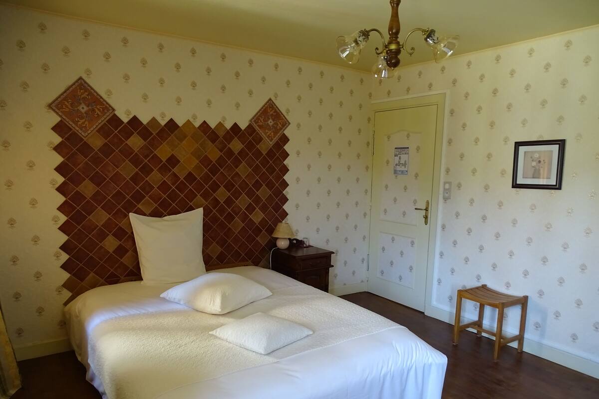 A bedroom with a large bed, a wooden headboard, a nightstand, a small stool, and a door with a poster.
