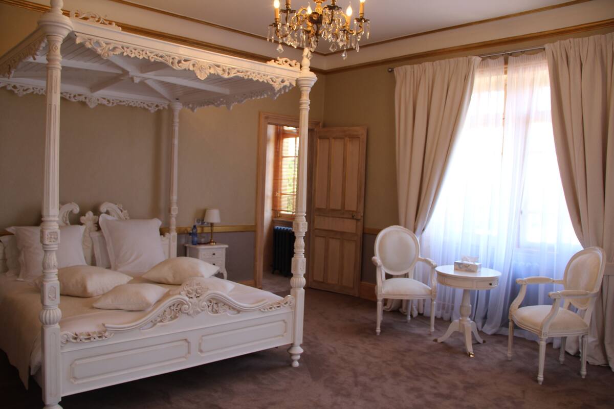 A four-poster bed with ornate detailing, a chandelier, and two white chairs in a room with a large window and a door in the background.