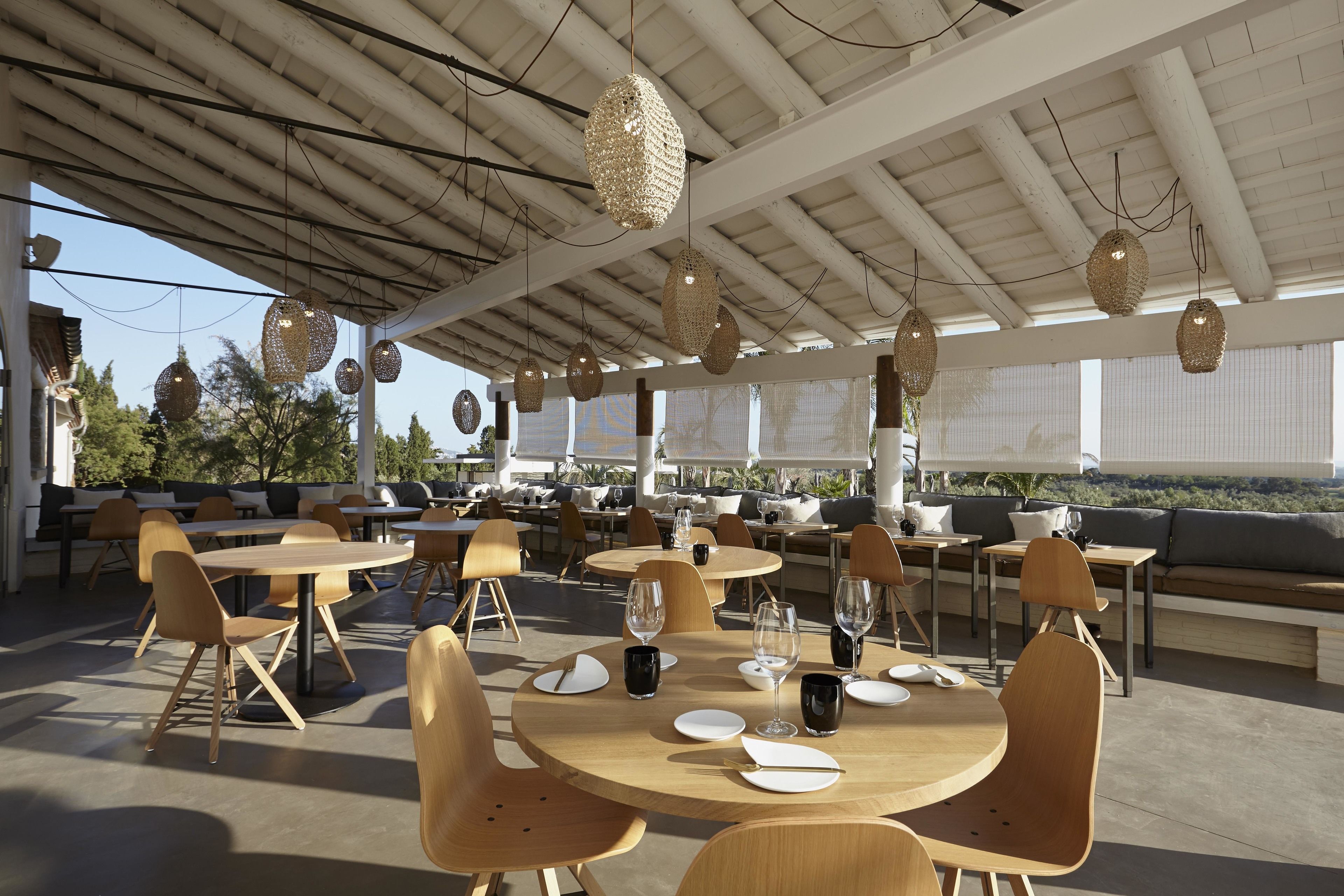 An outdoor dining area with round tables and chairs, hanging wicker lamps, and a view of the landscape.