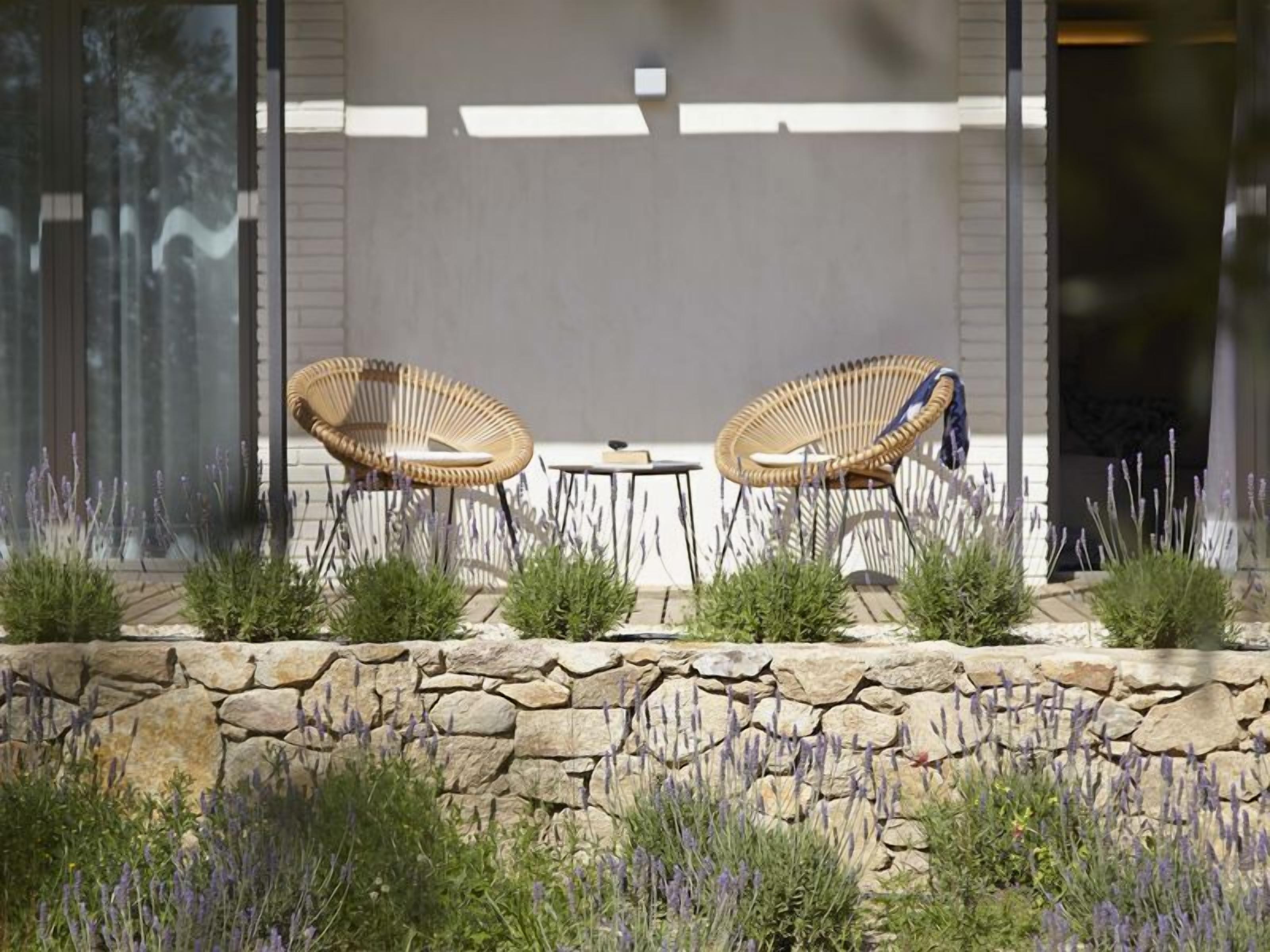 Two wicker chairs and a small table outside a building with a stone wall and lavender plants.