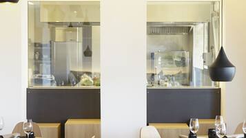 Un comedor moderno con mesas y sillas de madera, lámparas colgantes negras y vistas a la cocina a través de un separador de vidrio.