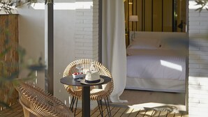 Un área de estar exterior acogedora con sillones de mimbre y una mesa, una cama blanca con cortina, y vista al interior a través de una puerta.