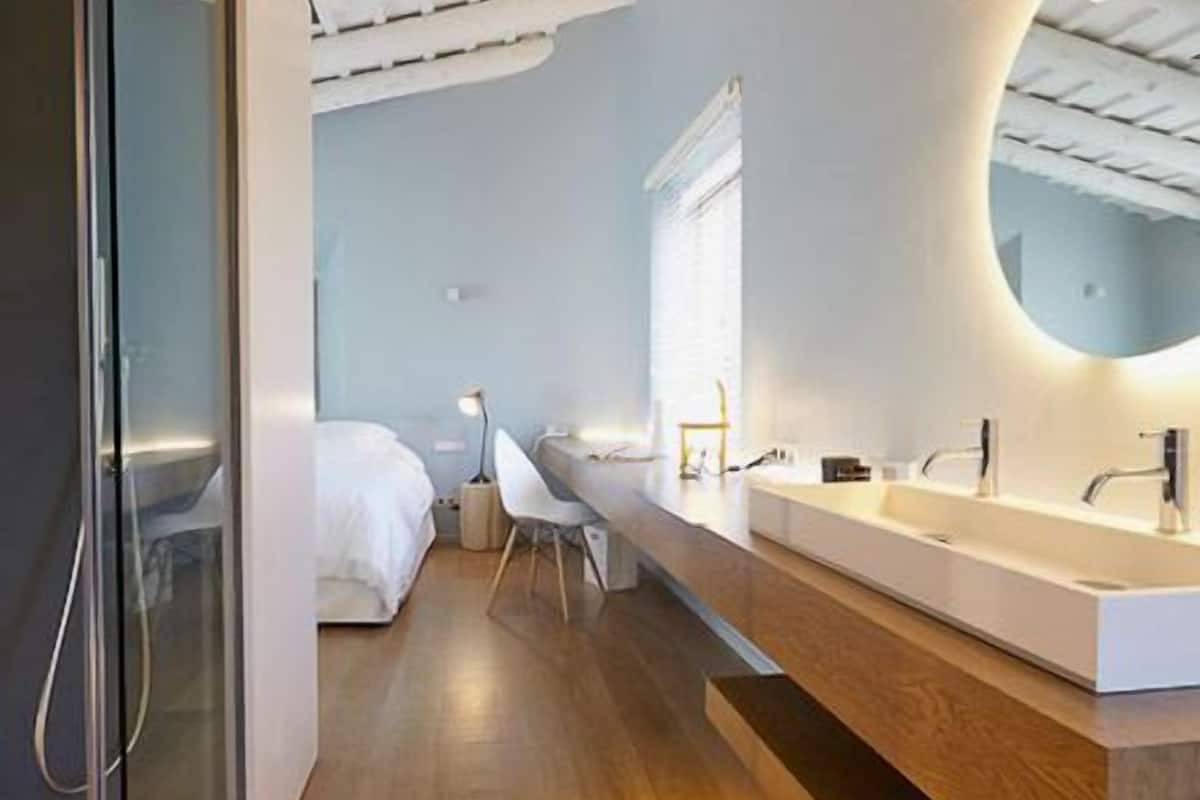 Une chambre d’hôtel moderne avec un plancher en bois, un lit blanc, un bureau avec une chaise, un grand miroir et une salle de bain blanche dotée d’un meuble-lavabo en bois.