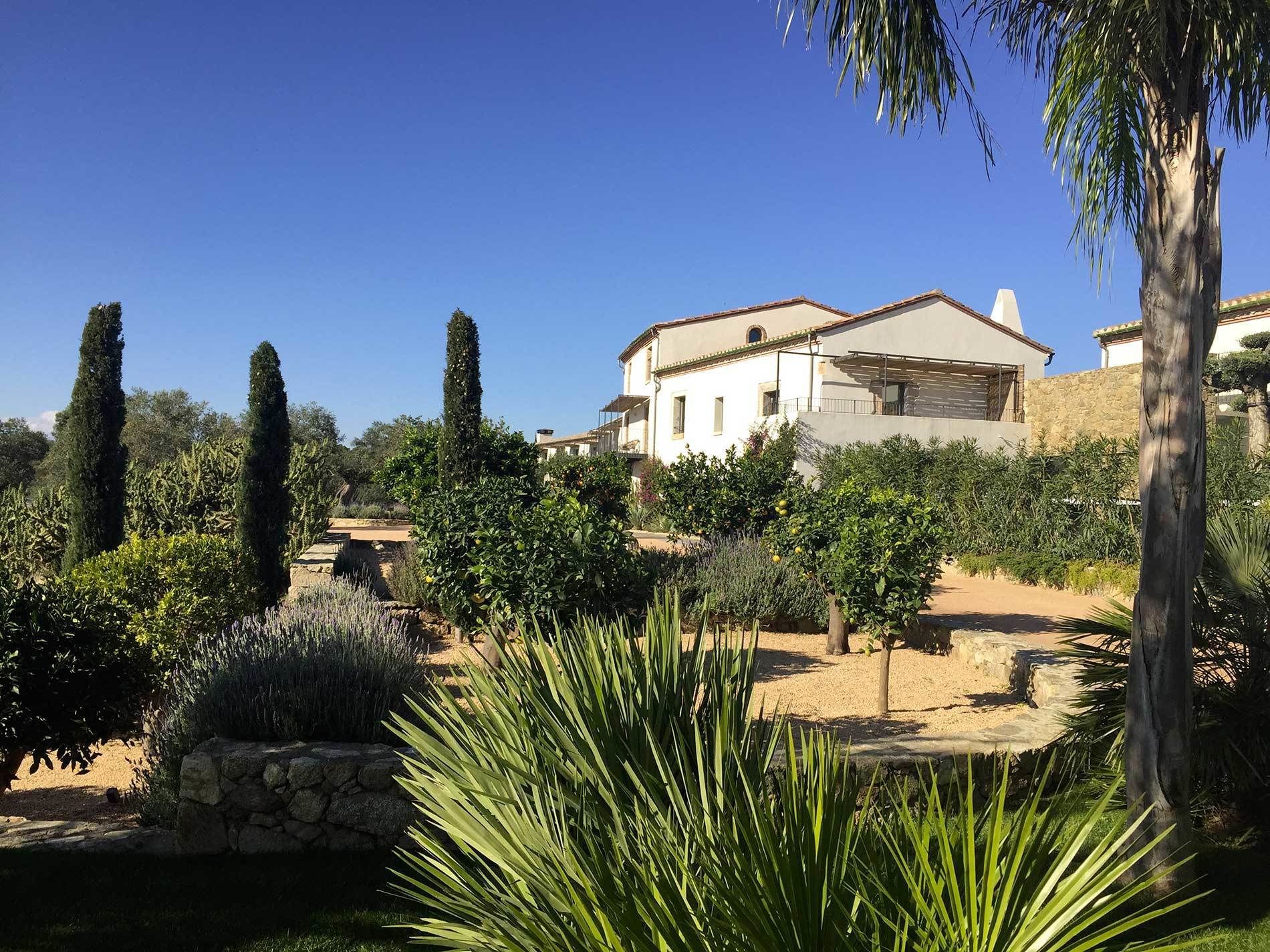 Una casa con un muro de piedra, un jardín con diversas plantas y un cielo azul despejado.