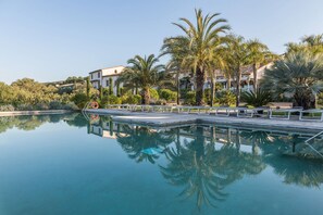 A clear swimming pool with a deck, surrounded by palm trees and a building with a tiled roof in the background. - Hotel Mas Lazuli (Pau)