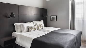 A modern bedroom with a dark wood headboard, a white bedspread, and a grey throw blanket. There are two bedside lamps and a framed picture on the wall.