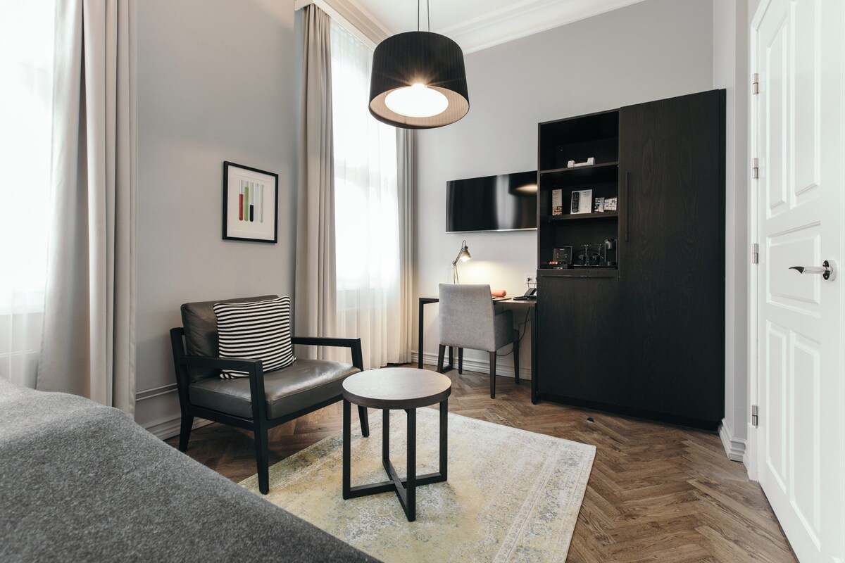 Ett modernt hotellrum med en soffa, ett litet runt bord, ett skrivbord med en stol, en TV som är monterad på väggen och en garderob.