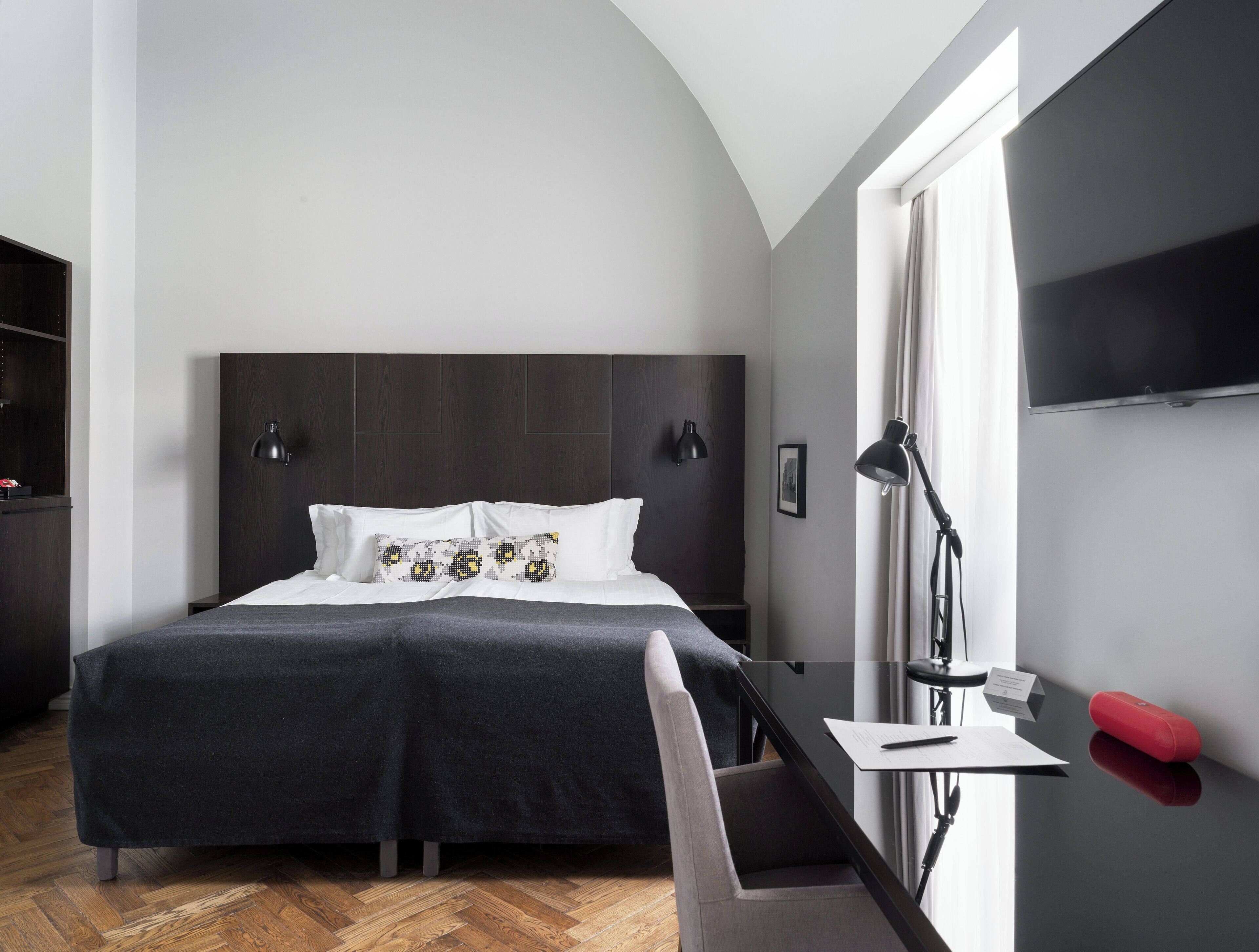 A modern hotel room with a large bed, a desk with a chair, a desk lamp, a red speaker, and a TV on the wall.