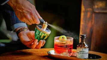 A person pouring a drink from a green shaker into a glass with a lime wedge. A bottle labeled "A POTEK'S BLACK DEATH NEGRON" is on a tray beside the glass.