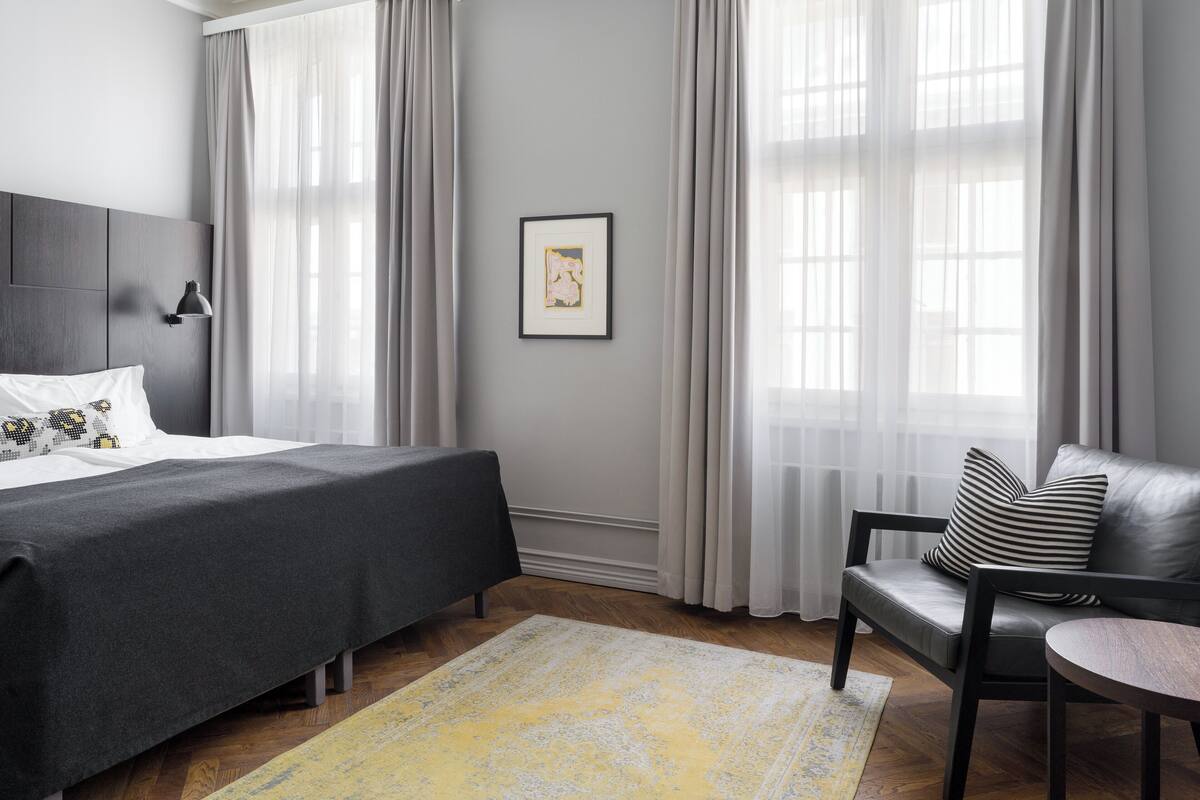 Ein modernes Hotelzimmer mit Bett, einem Sessel, einem kleinen Tisch und großen Fenstern mit Vorhängen.