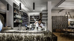 Un espace bar doté d’un comptoir en marbre, de plusieurs étagères remplies de bouteilles d’alcool et d’un barman préparant des cocktails.