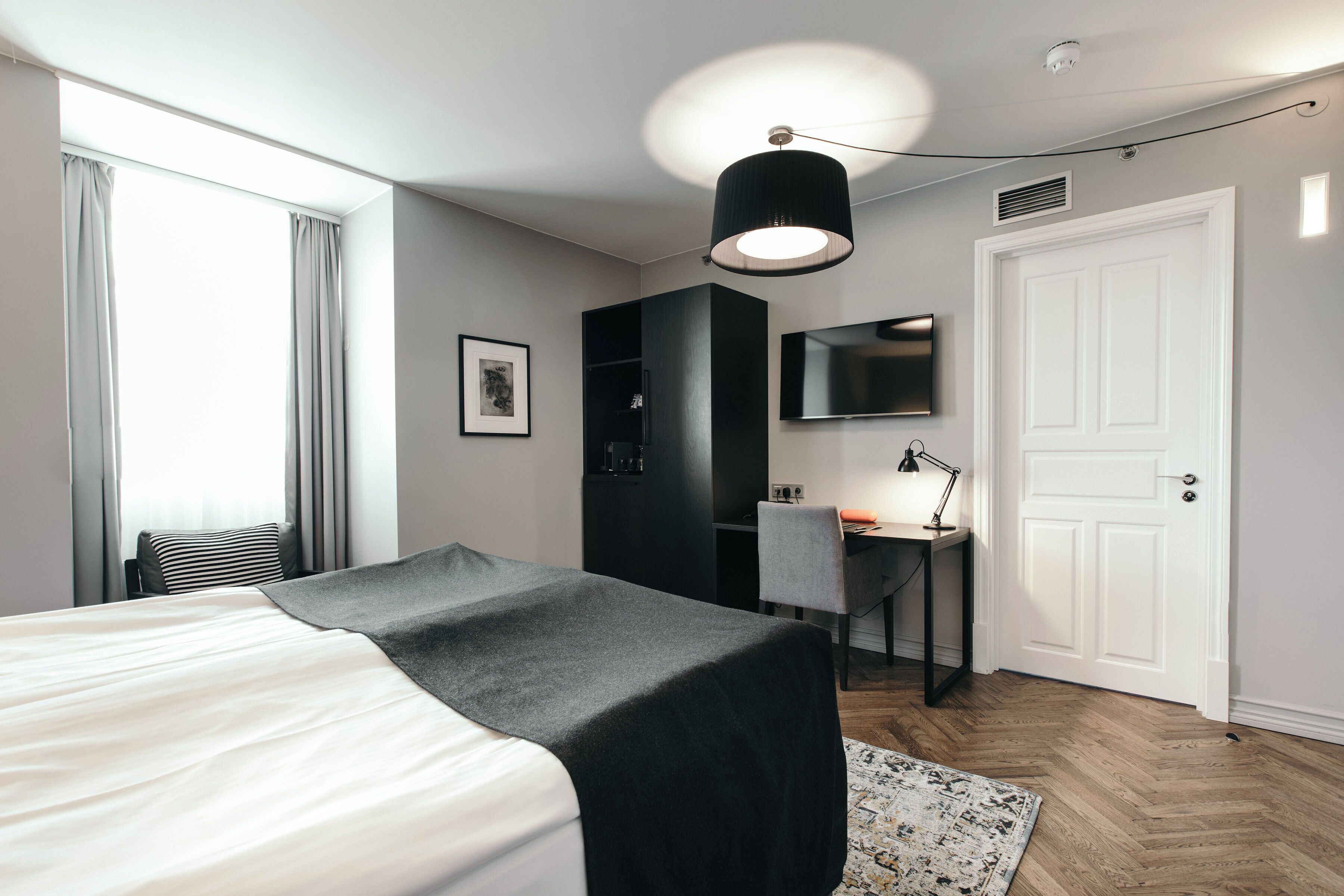 A modern hotel room with a large bed, a desk, a chair, a wardrobe, a mirror, and a door.