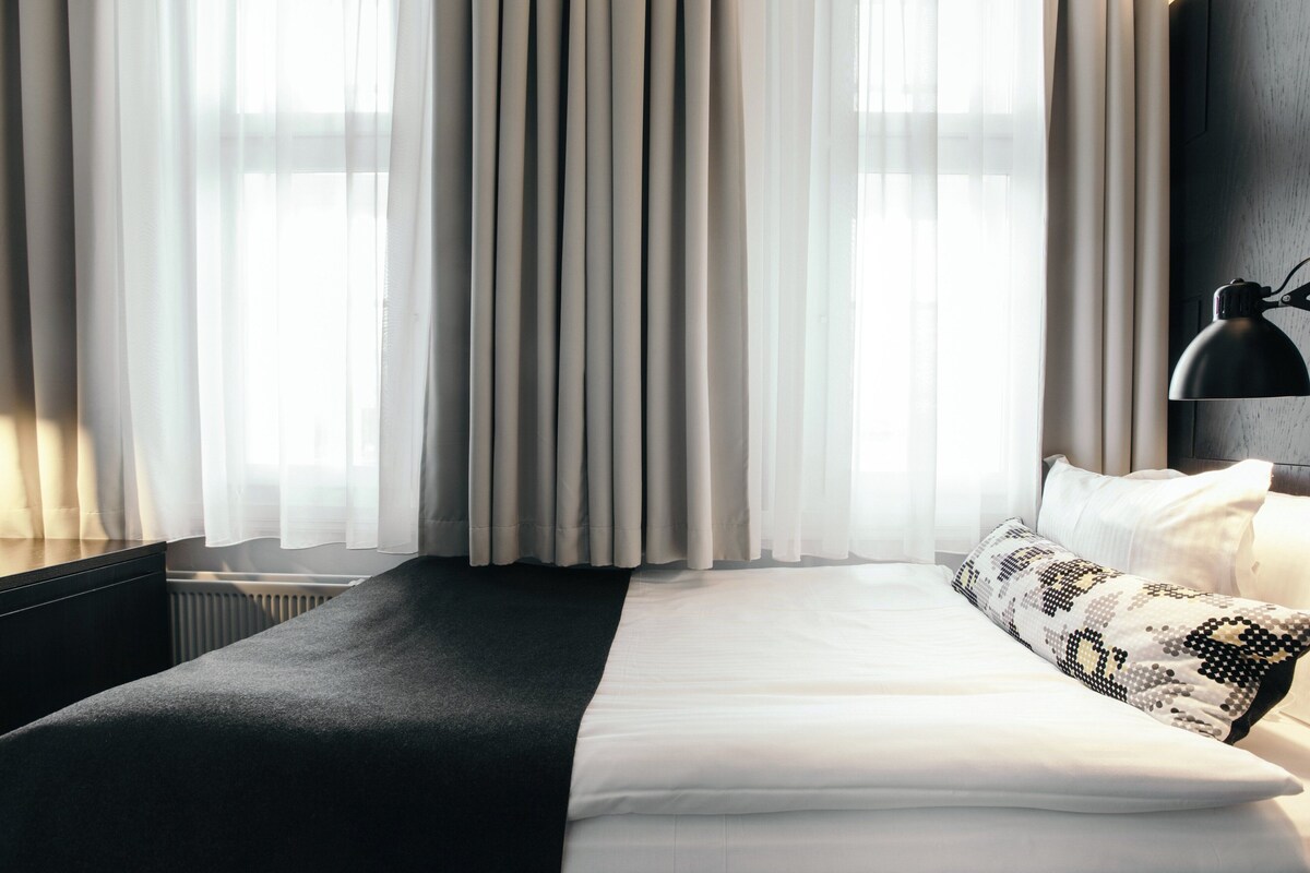 Ett hotellrum med en stor säng, ett svart sängbord, en lampa och tunna, genomskinliga gardiner.