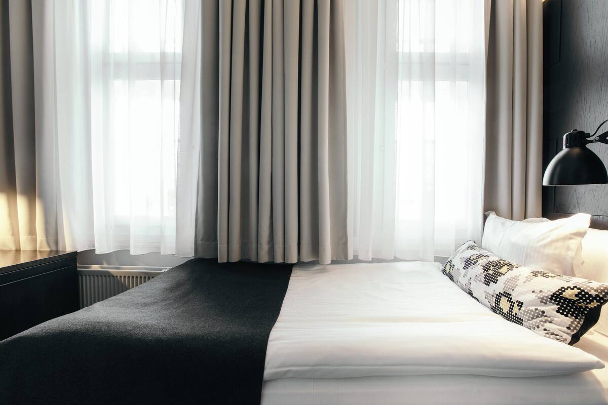 Ein Hotelzimmer mit einem großen Bett, einem schwarzen Nachttisch, einer Lampe und durchsichtigen Vorhängen.