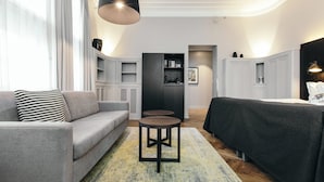 Un salon moderne avec un canapé gris, une table basse ronde et une table à manger noire recouverte d’une nappe foncée. On y trouve également un coin cuisine avec des placards et un luminaire suspendu.