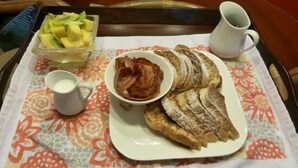 A breakfast tray with sliced bread, bacon, a bowl of fruit, and a cup of milk on a patterned tablecloth.