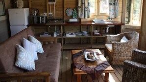A cozy cabin interior with a wooden dining table, wicker chairs, a brown sofa with patterned cushions, and a kitchen area with a refrigerator, toaster, and window with curtains. - Gourits River Guest Farm (Albertinia)