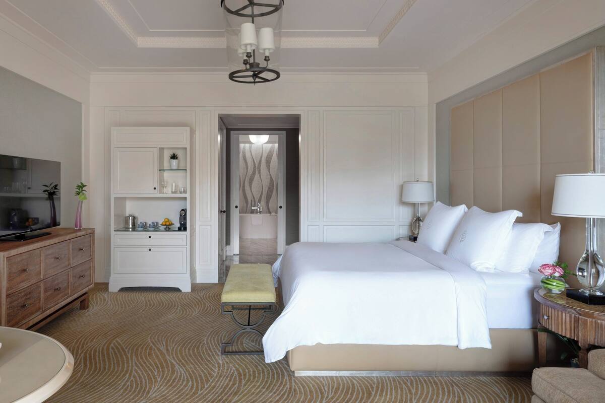 A hotel room with a bed, a TV, a desk, a chair, a bedside table, and a bathroom visible through an open door.