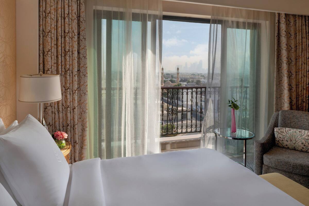 A hotel room with a large bed, a sitting area with a chair, a small table with a vase, and a view of a cityscape through sheer curtains.